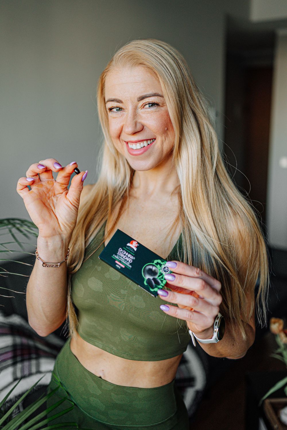 Smiling blonde woman in green crop top holding a dark green product card with both hands.