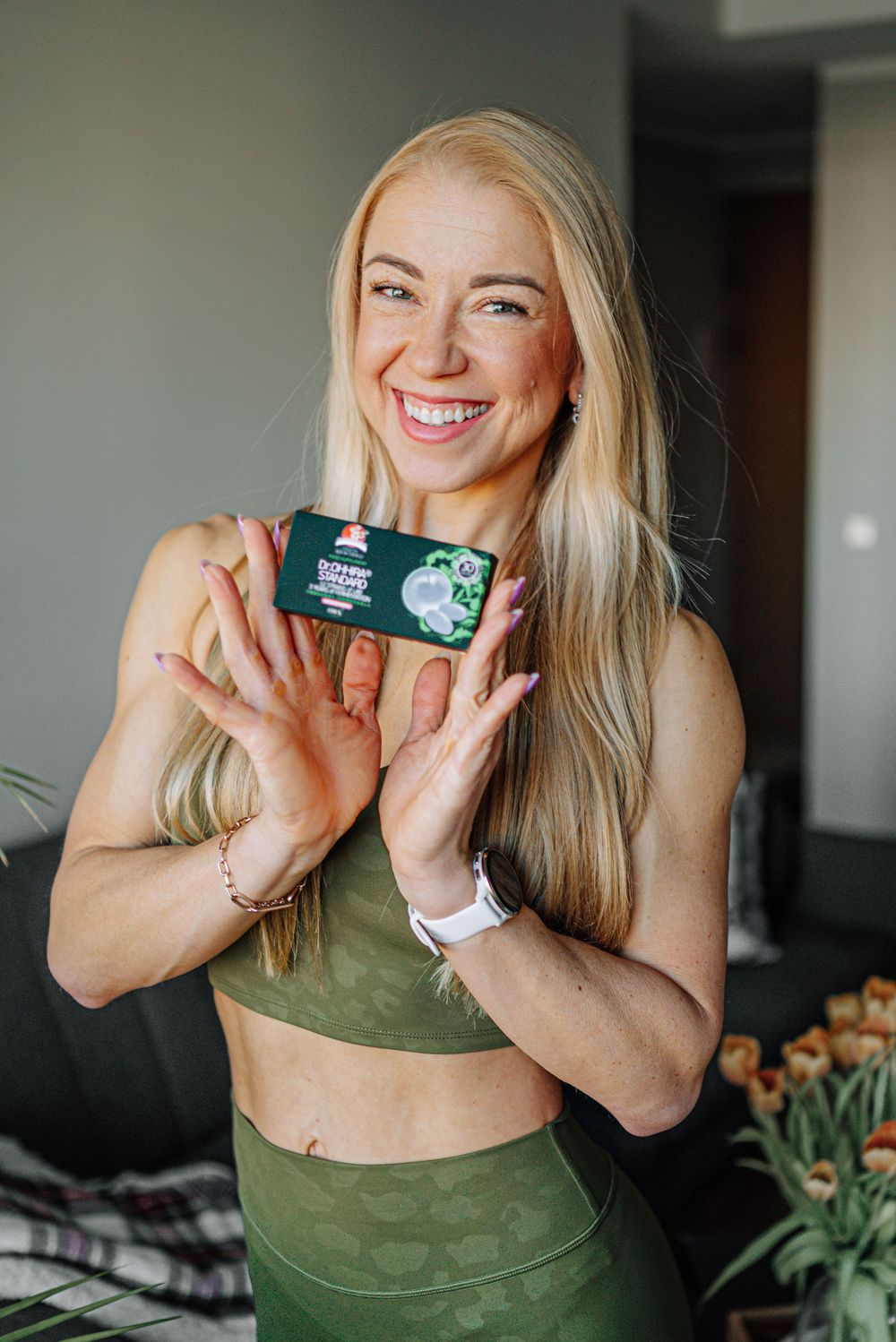 Smiling blonde woman in green athletic wear holding a dark green product box with both hands.