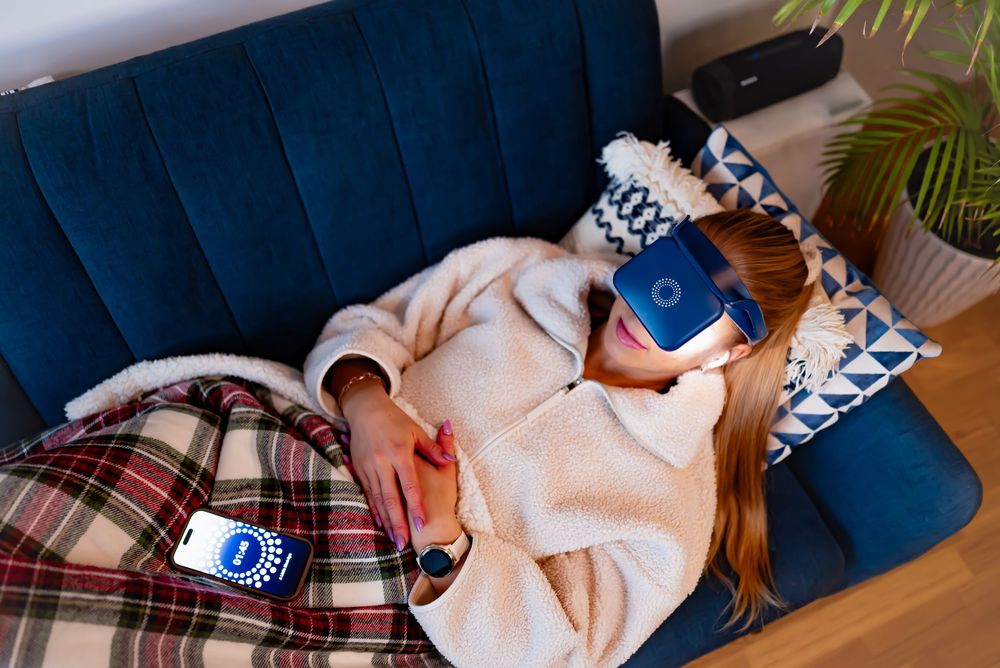 Woman lying on blue sofa wearing VR headset and white robe, covered with plaid blanket, holding smartphone.