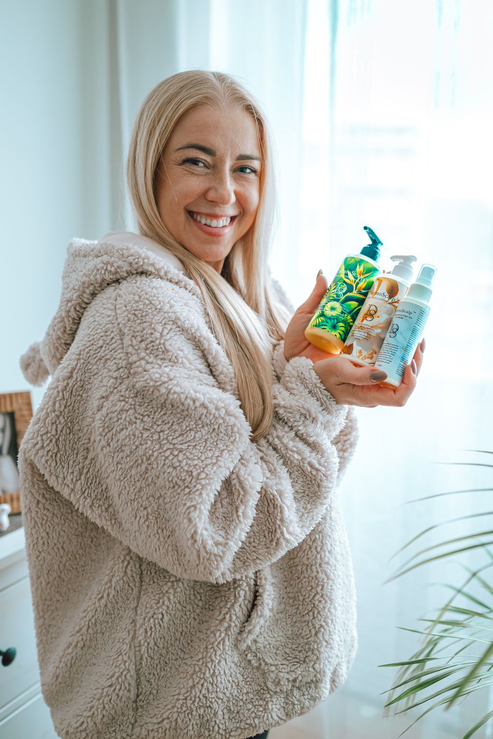 Woman in cozy teddy bear coat holds skincare products while posing against bright window background.