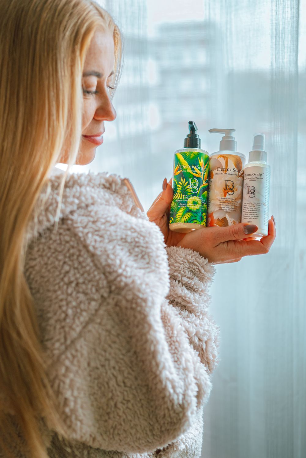 Woman in cozy white sweater holding skincare products and examining beauty product bottles near window.