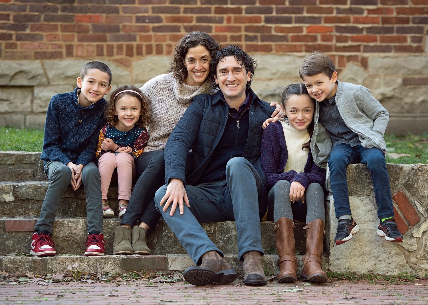 Happy family of six smiling together on stone steps outdoors near a brick wall in autumn.