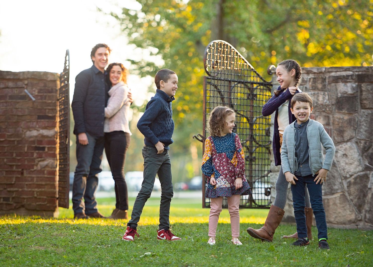 Family of six posing outdoors near ornate gate, children playing with badminton racket on green lawn with autumn trees.