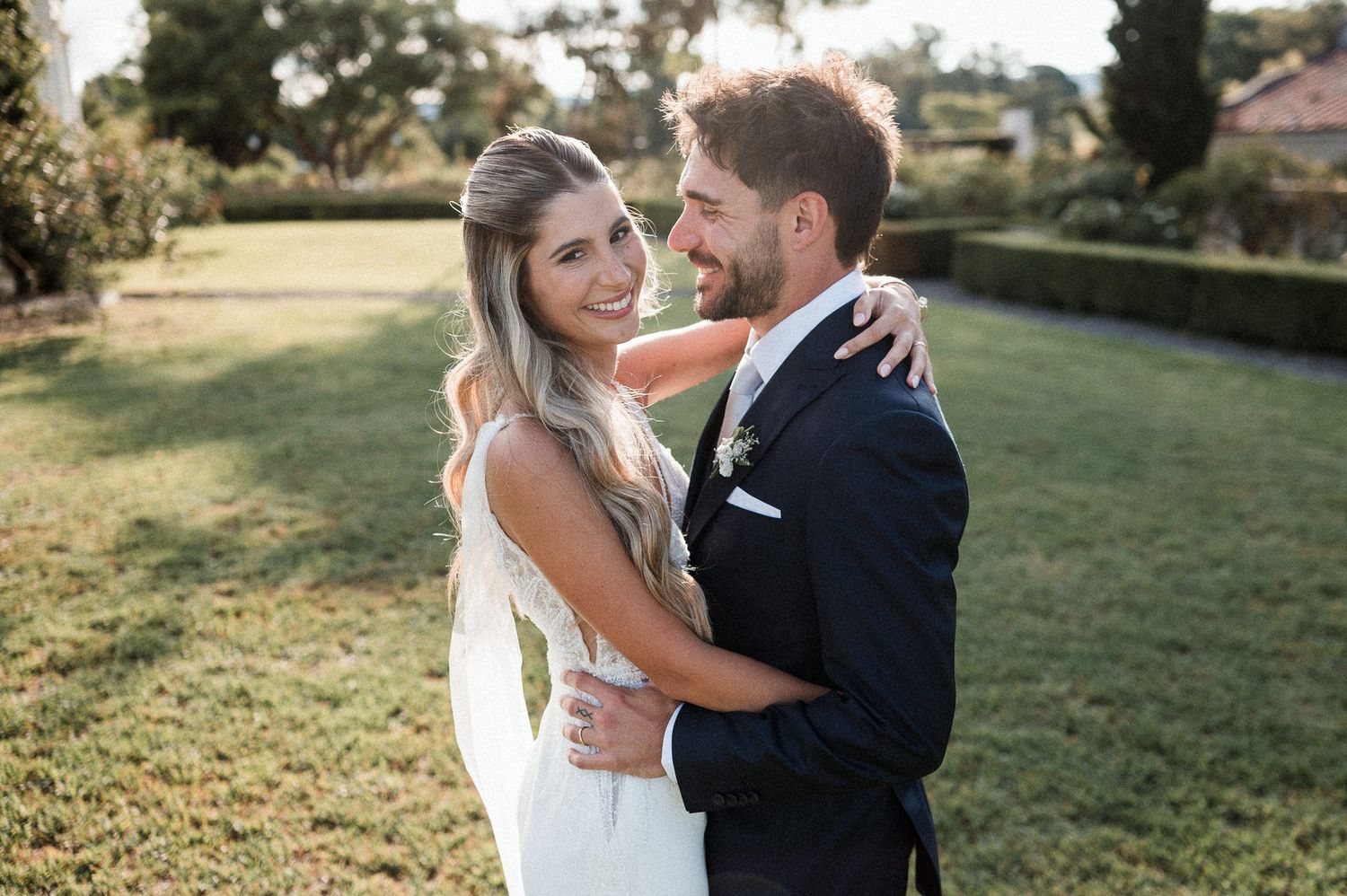 Newlyweds embracing on a sunny lawn with elegant wedding attire.