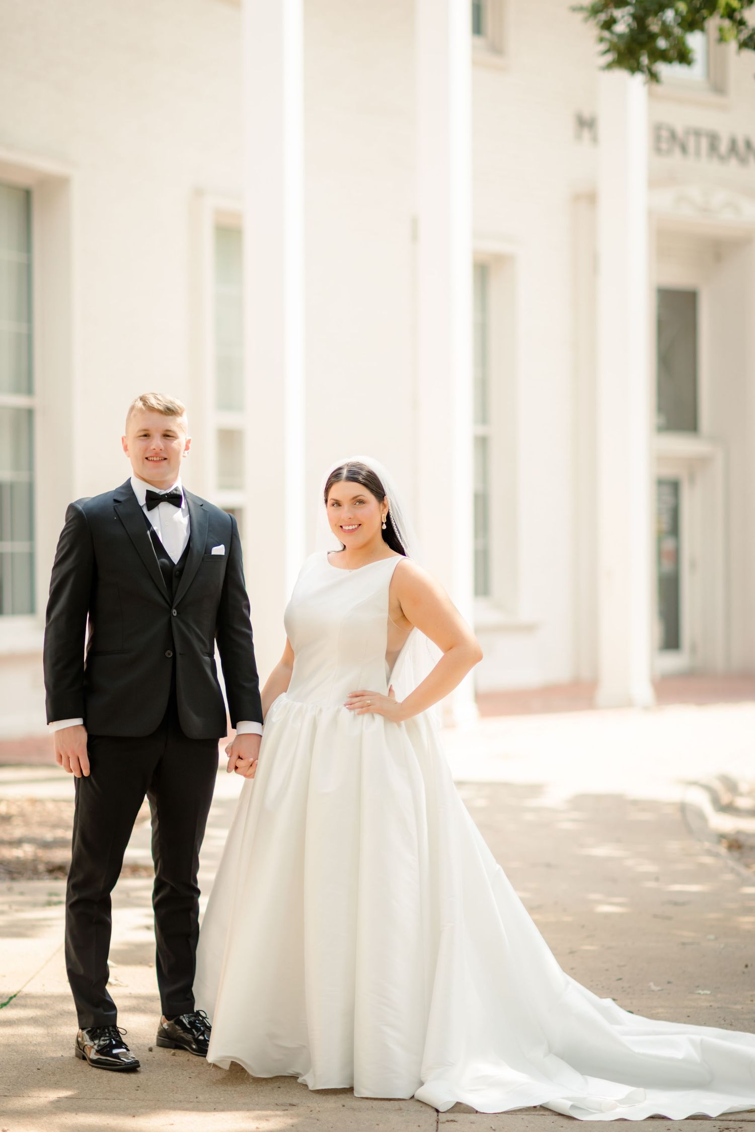 A bride in a flowing white gown and groom in black tuxedo pose together in front of a bright colonnade.