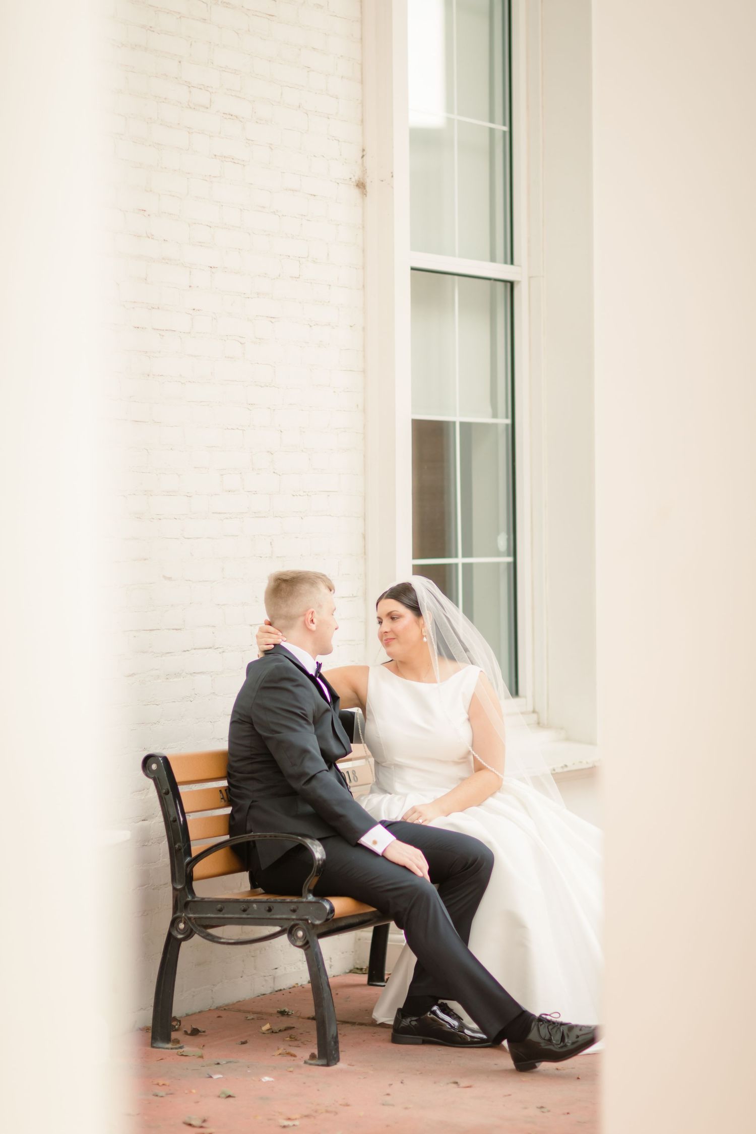 A bride and groom share a quiet moment while sitting on wooden benches outside a white building.