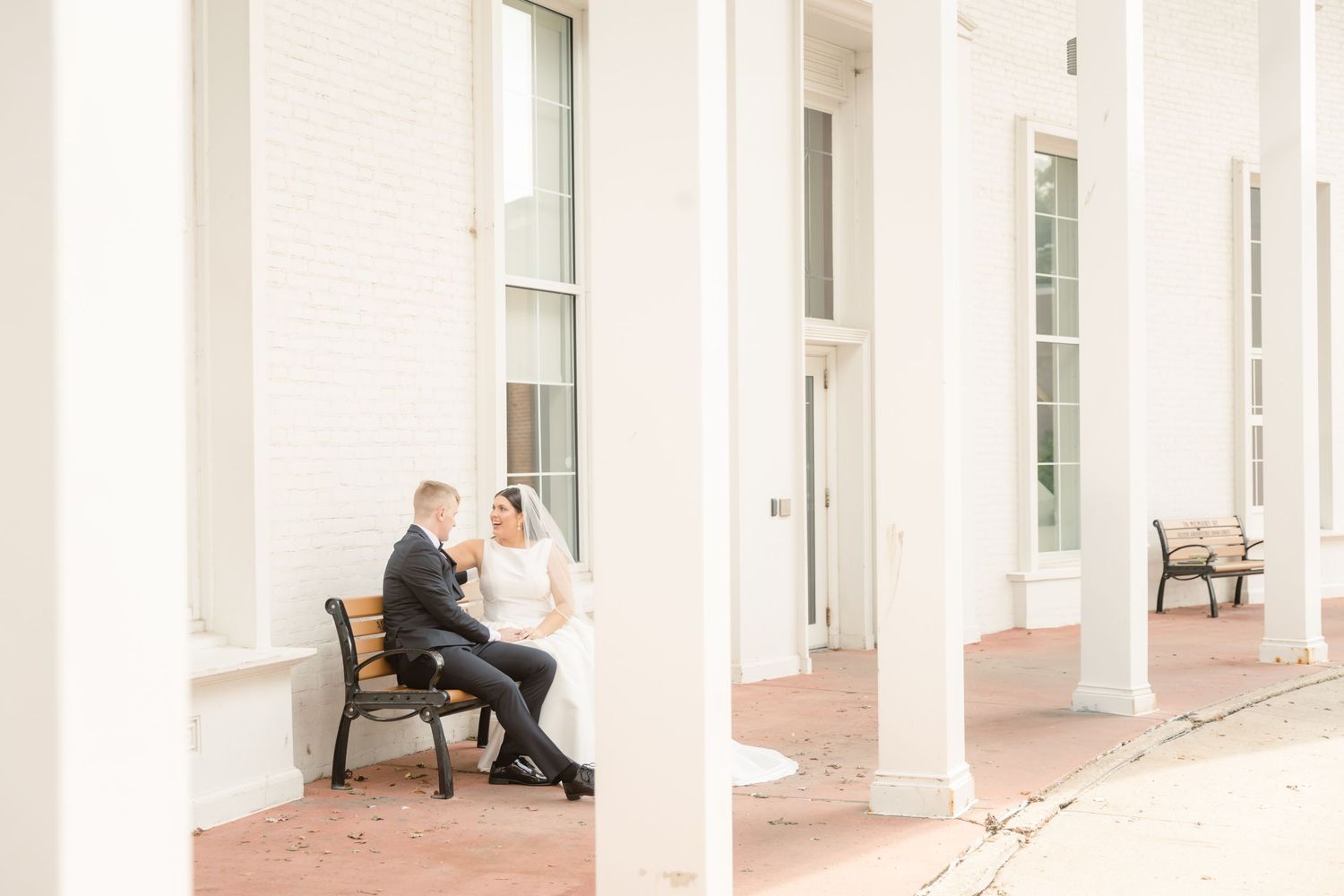 A couple sits together on a wooden bench between white columns on an elegant porch.