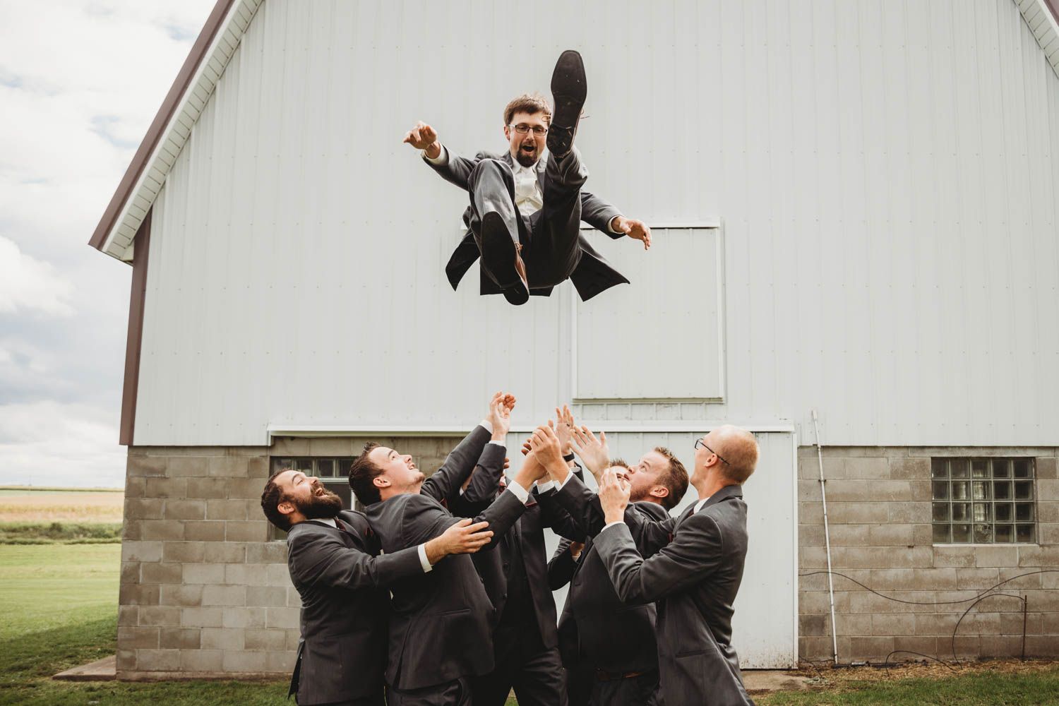 Wedding party members leap joyfully in the air outside a rustic barn setting in a synchronized group photo.