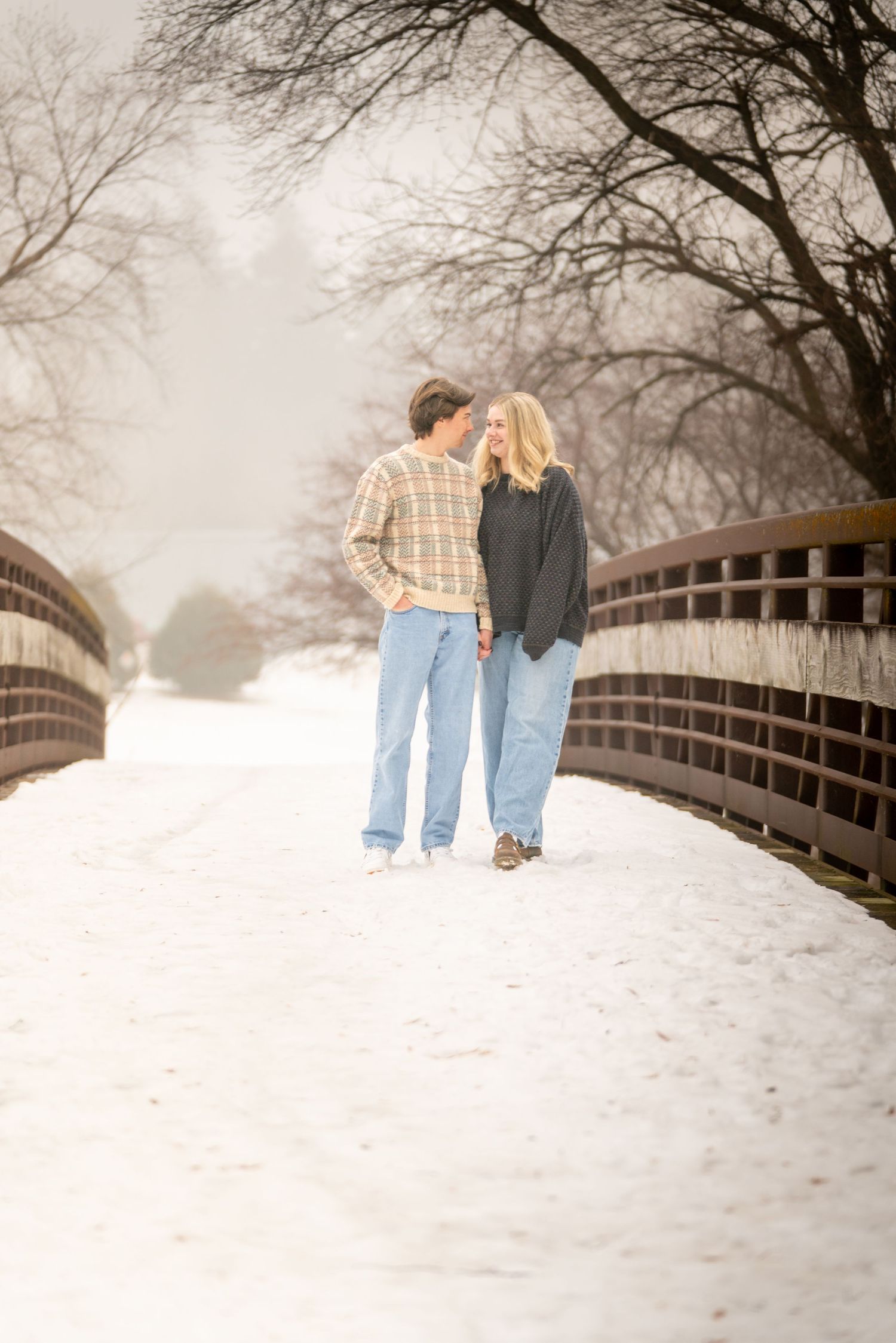 Couple holding hands on a snow-covered bridge in winter, surrounded by bare trees and a misty landscape.