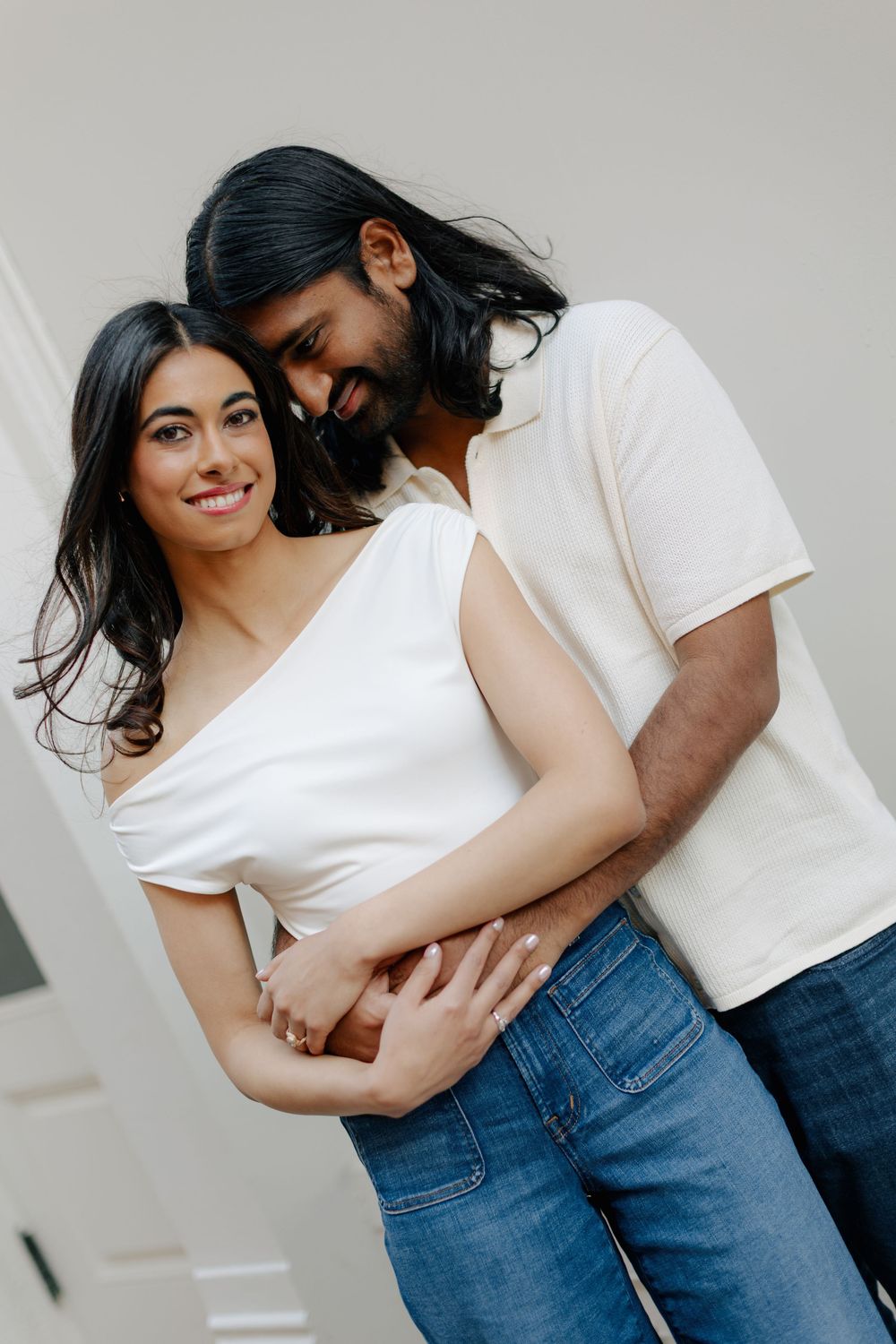 A romantic couple in white tops and jeans embrace intimately against a light background.