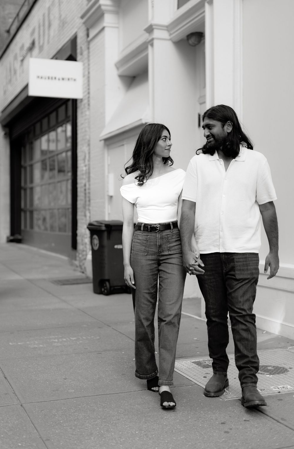 Two people in white shirts and jeans walking together on a city sidewalk in black and white.