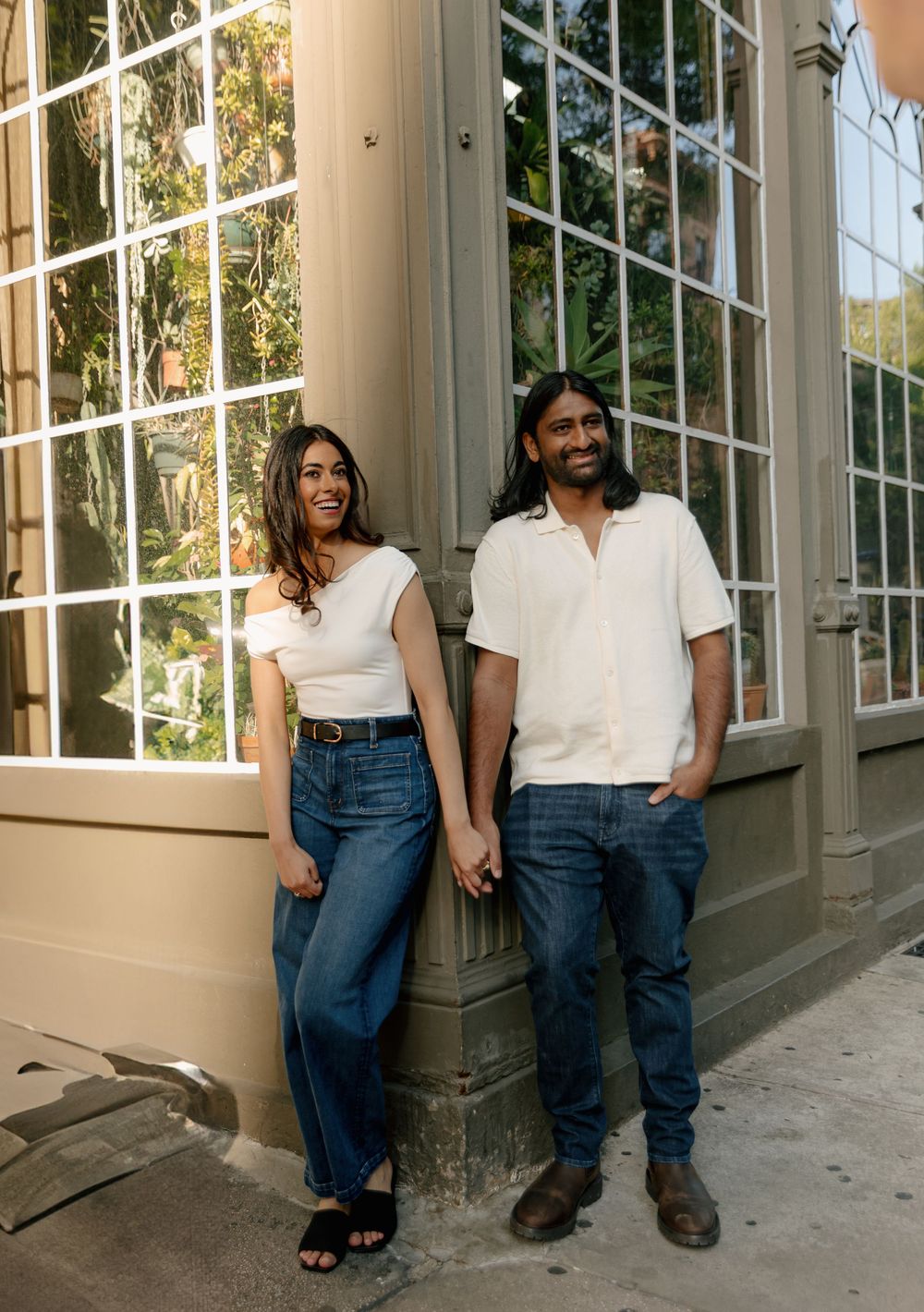 Couple in casual white tops and blue jeans standing together by sunny windows.