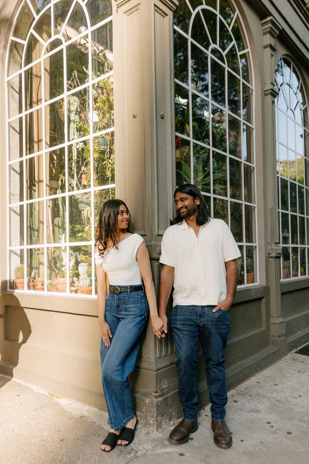 A couple in matching white tops and jeans poses against a building exterior with large windows.