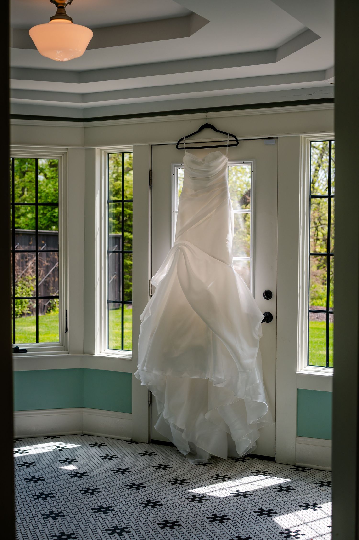 A white wedding dress hangs in a sunny room with turquoise walls, black and white tiled floor, and bay windows.