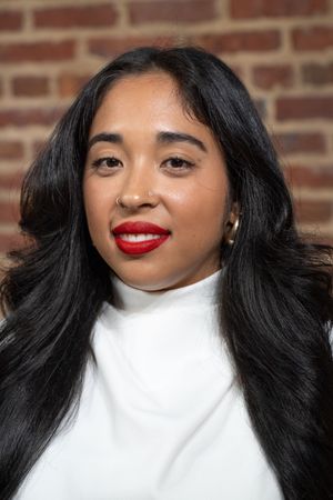 A person with long black hair and bright red lipstick poses in front of a brick wall wearing a white top.