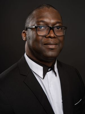 Professional headshot of person in black suit and white shirt against dark background wearing glasses.