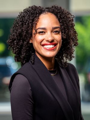 Professional headshot portrait of someone in a black blazer with curly hair and bright smile against blurred background.