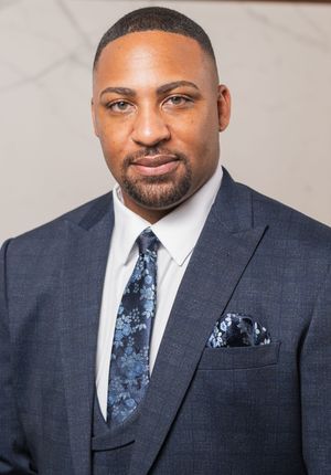 Professional headshot of someone wearing a dark suit with a blue patterned tie and matching pocket square.