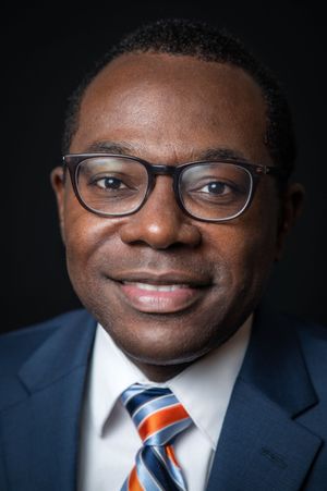 Professional headshot of a person wearing glasses, a navy suit and striped orange tie against a dark background.