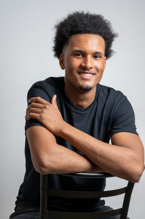 A person in a black t-shirt poses with crossed arms while sitting on a chair against a light background.