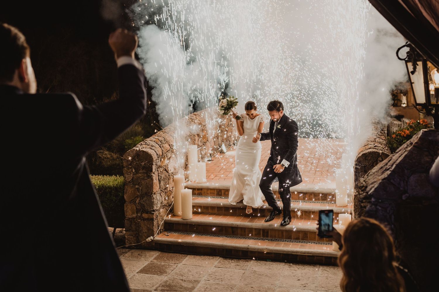 Newlywed couple descends stone steps amid dramatic smoke and sparkler effects at evening wedding celebration.