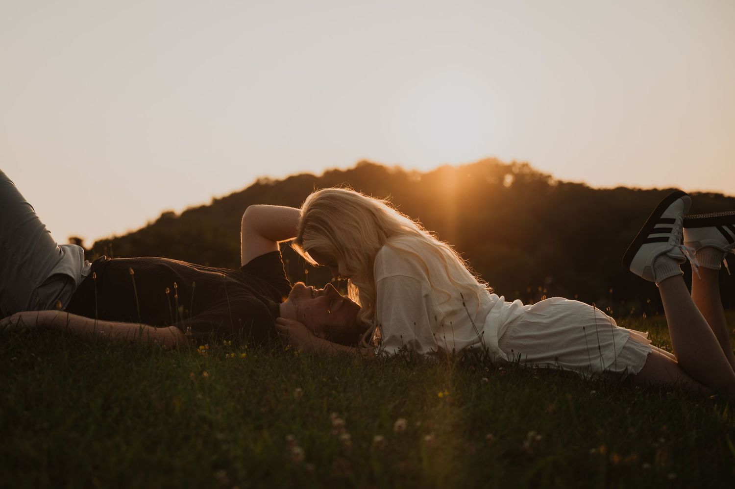Two people lying in grass at sunset with golden sunlight streaming through creating a warm, romantic atmosphere.