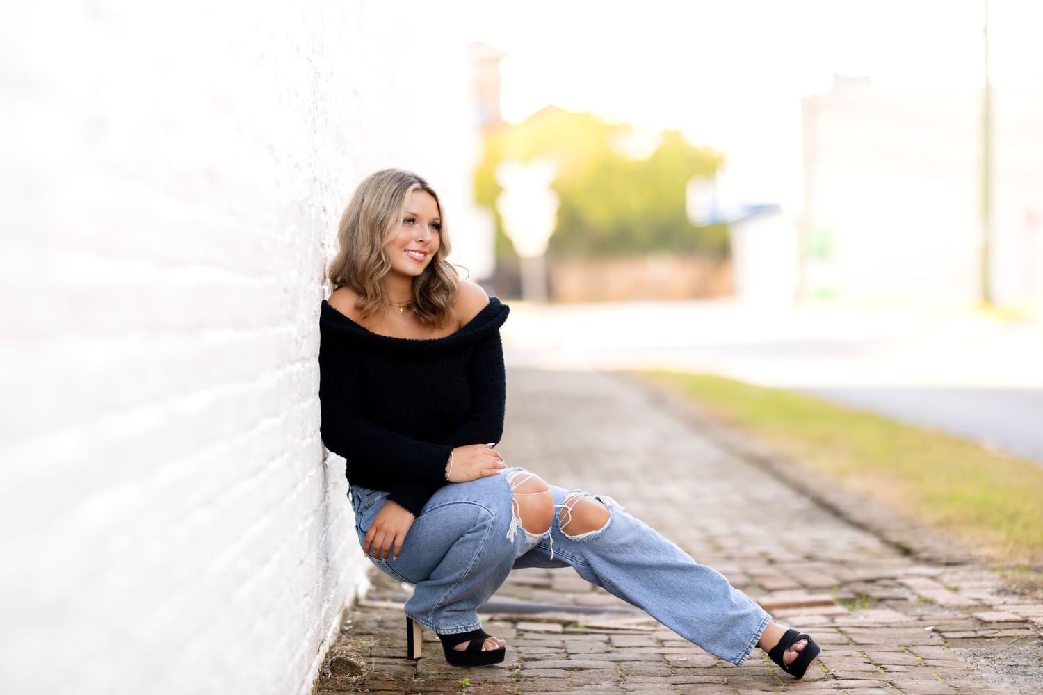 Casual portrait of someone in ripped jeans and black sweater sitting against white wall during golden hour.