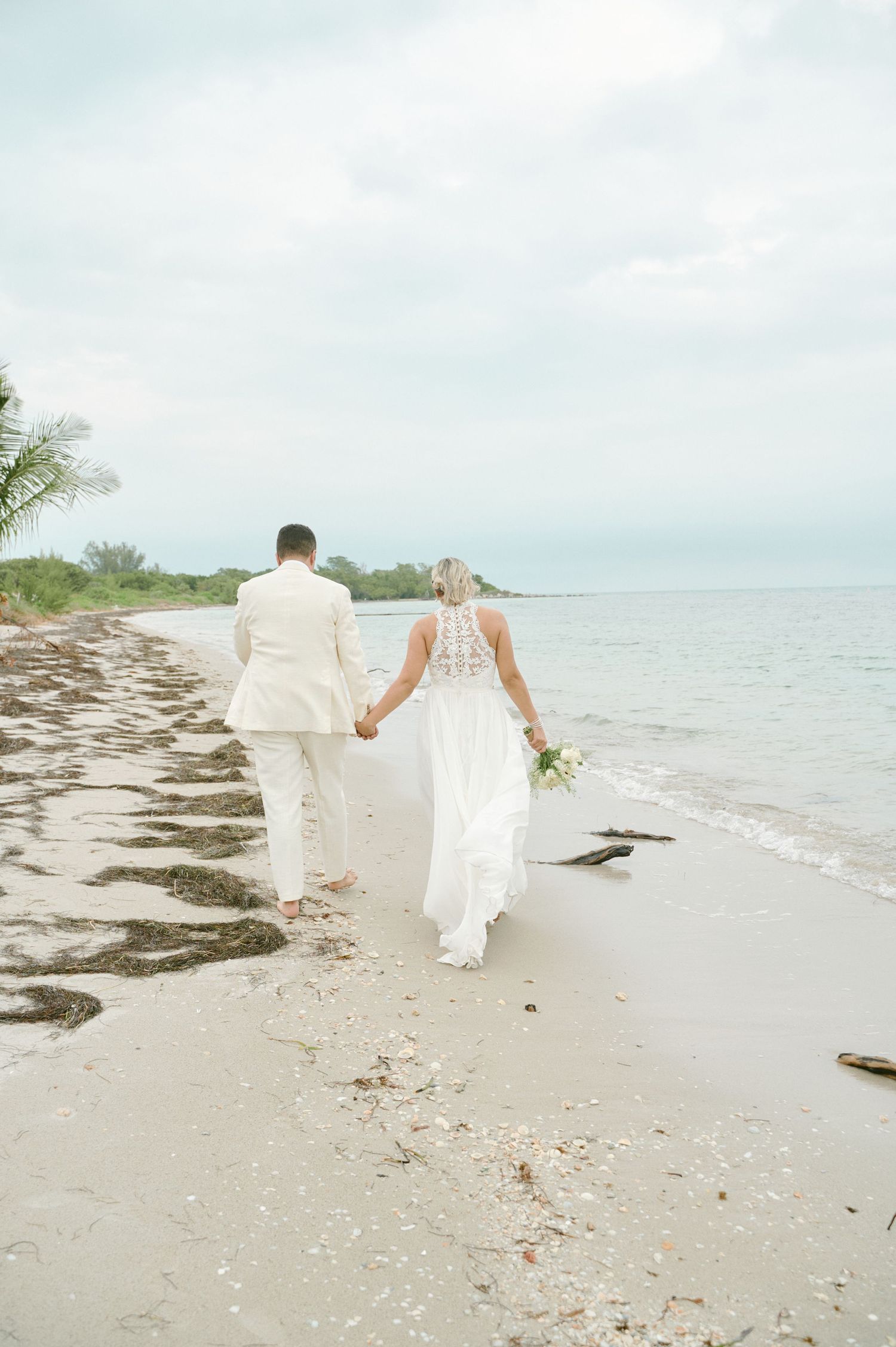 Couple walking hand in hand along beach shore in elegant wedding attire.