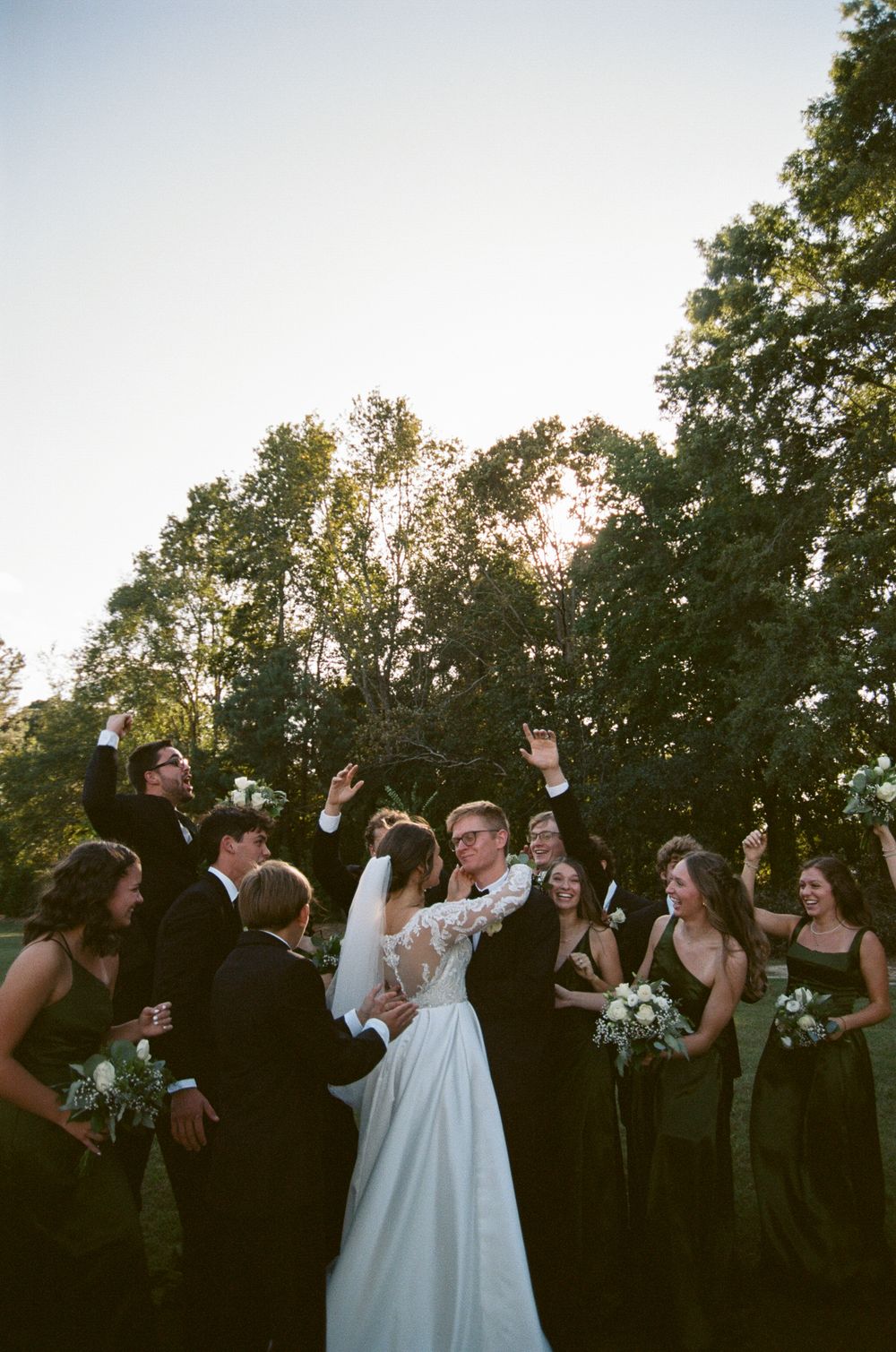 Wedding party celebrates outdoors at sunset with joyful poses and raised arms against a backdrop of trees.