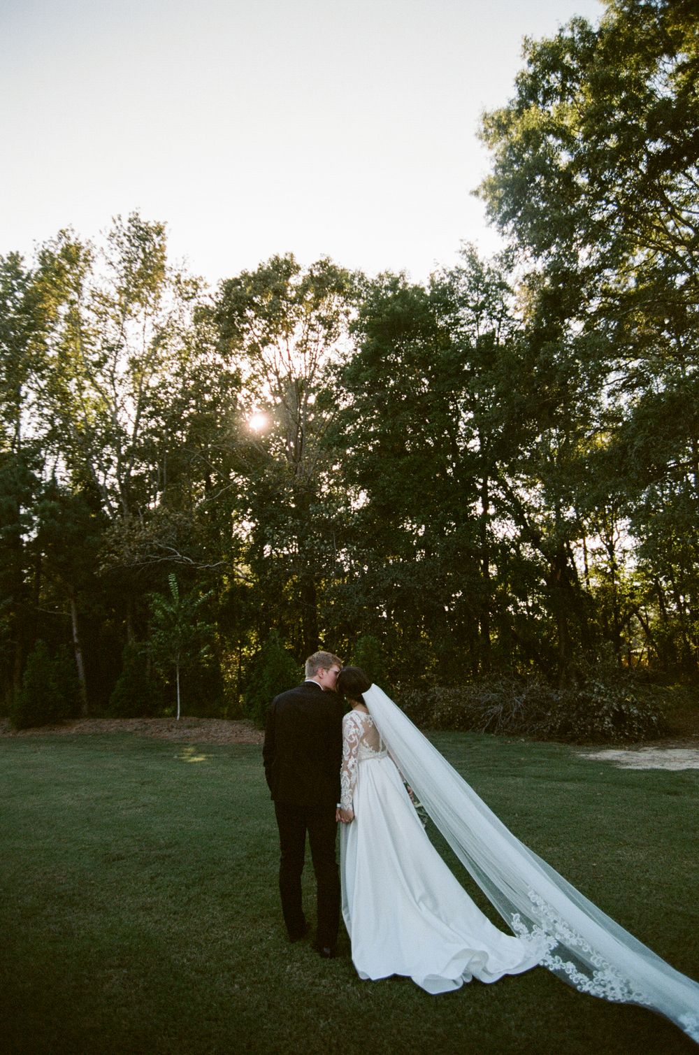 A wedding couple stands in a scenic outdoor setting at sunset with trees in the background.