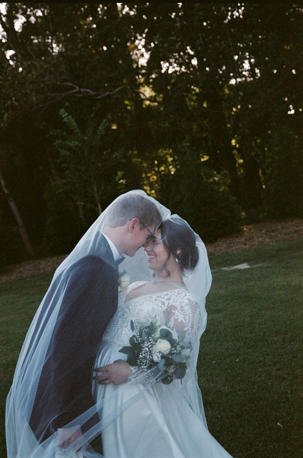 A romantic wedding couple shares a tender moment under a flowing veil during their outdoor sunset photo session.