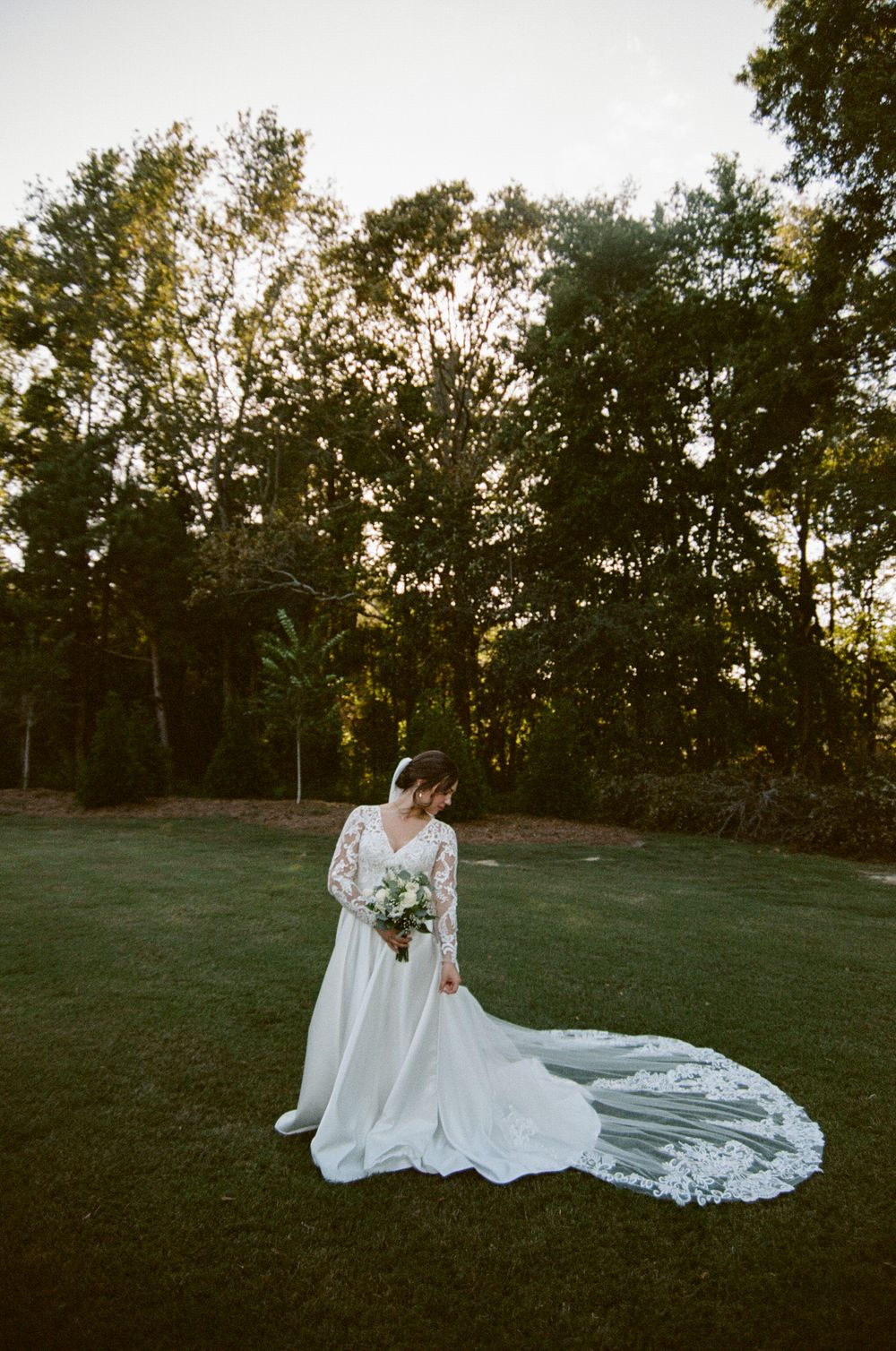 A bride in a long-sleeved white wedding gown with a flowing lace train sits gracefully on a grassy hillside at sunset.