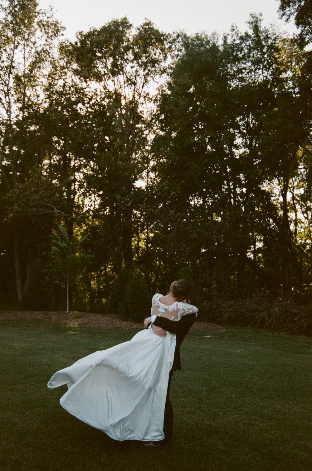A person in a flowing white dress twirls gracefully in a grassy field at dusk with trees in the background.