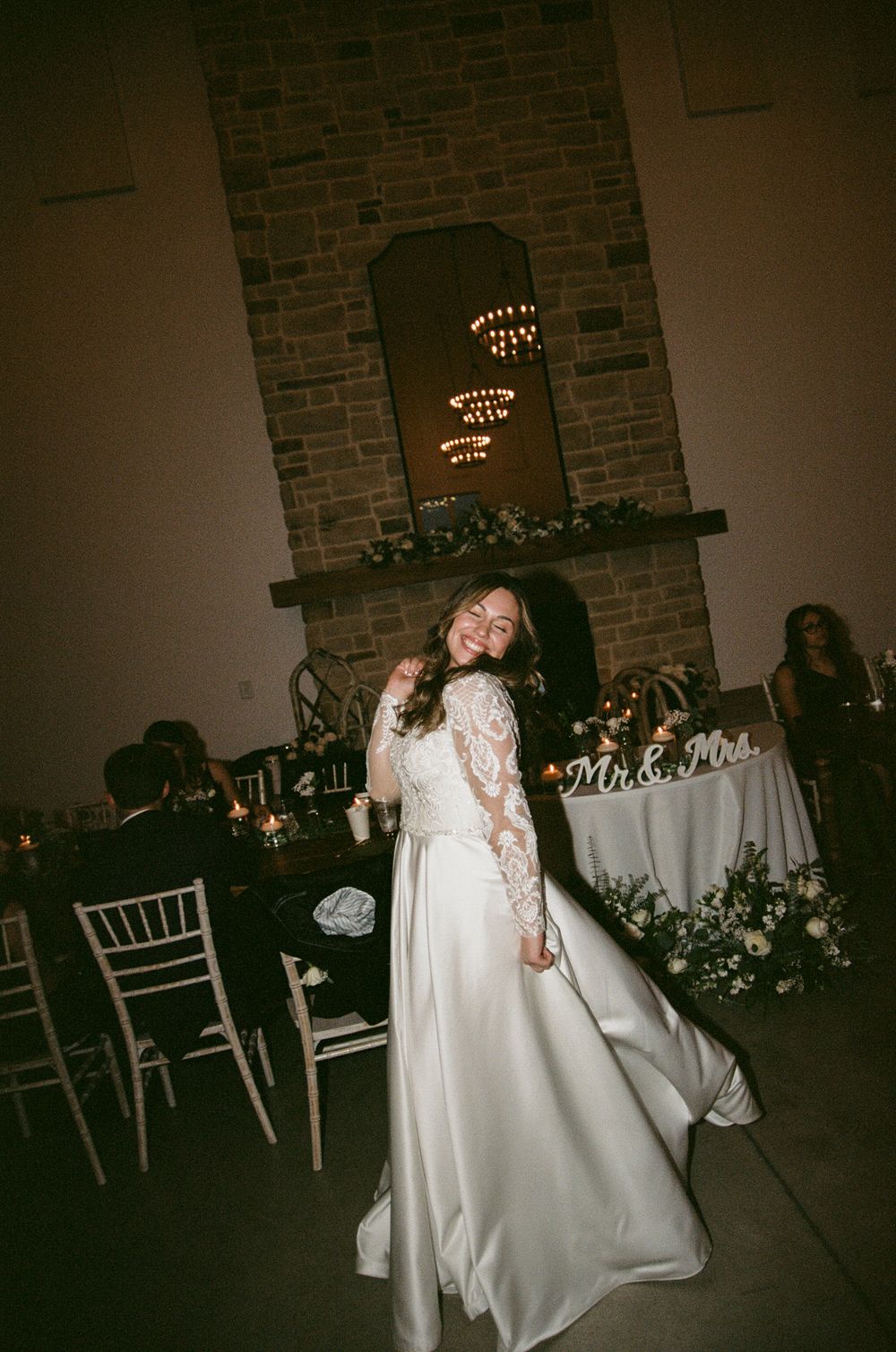 A bride in a flowing white wedding gown twirls and dances in an elegant reception venue with brick walls and tables.