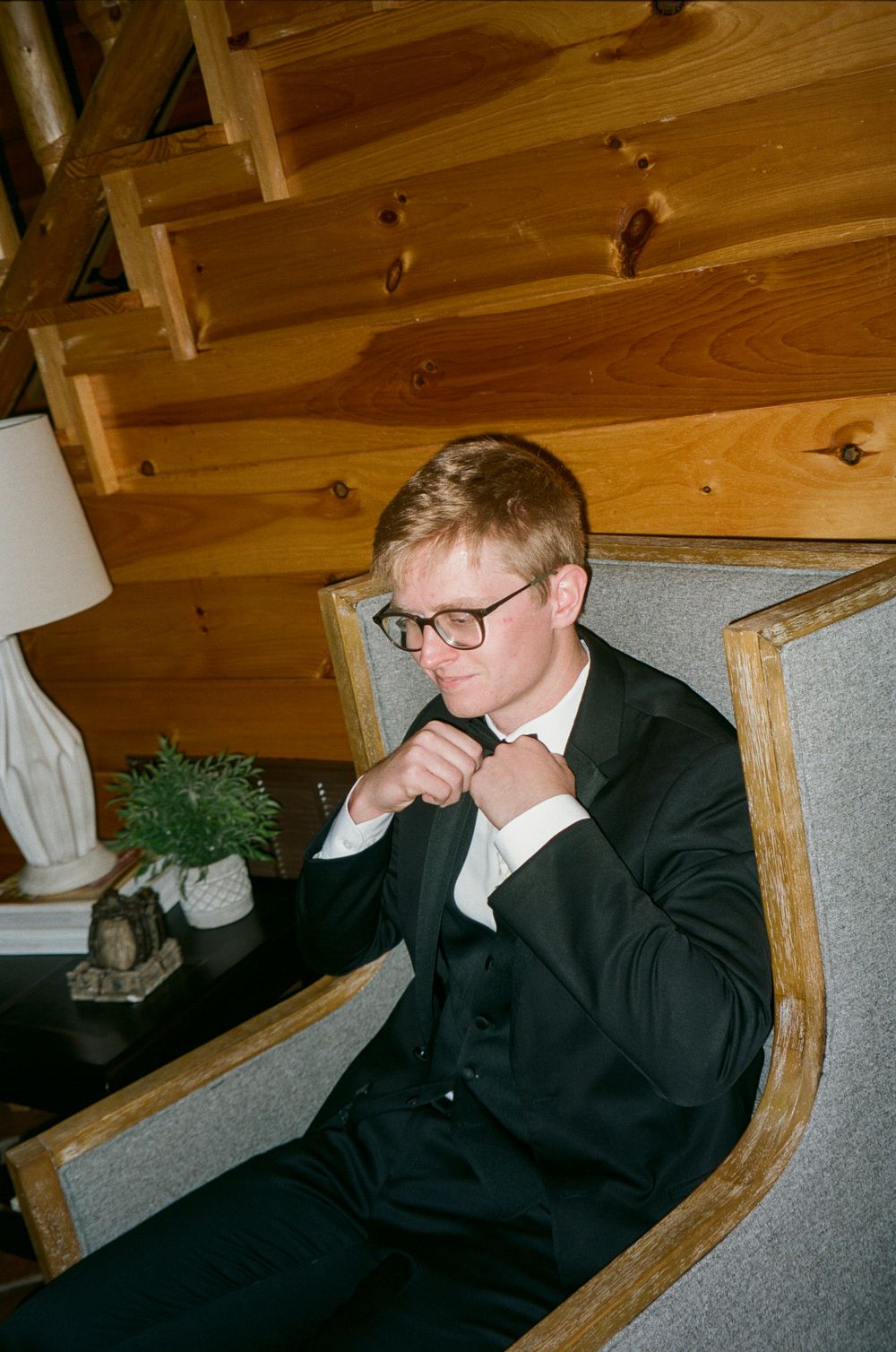 A person in a dark suit and glasses sits thoughtfully in a wooden interior with rustic decor.
