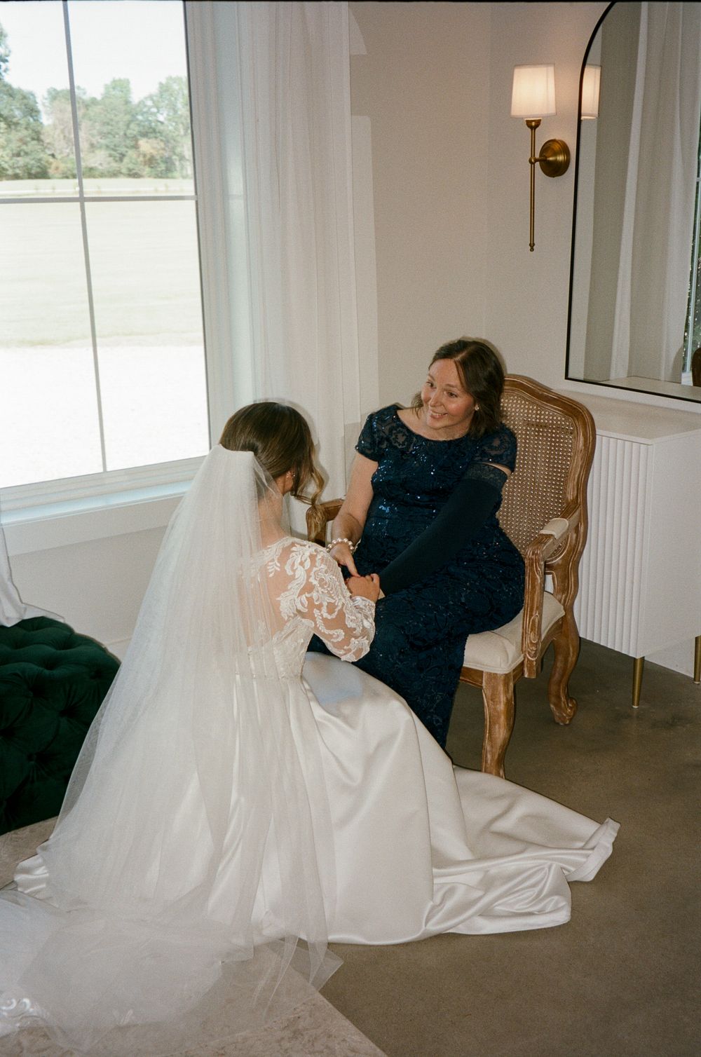 Wedding dress fitting scene in a bright sunlit room with white walls and wooden chairs.