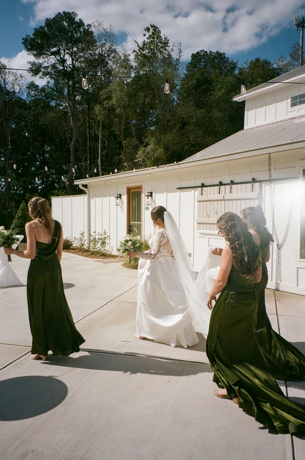 Wedding party in dark dresses and white gown walking along an outdoor white building pathway on a sunny day.