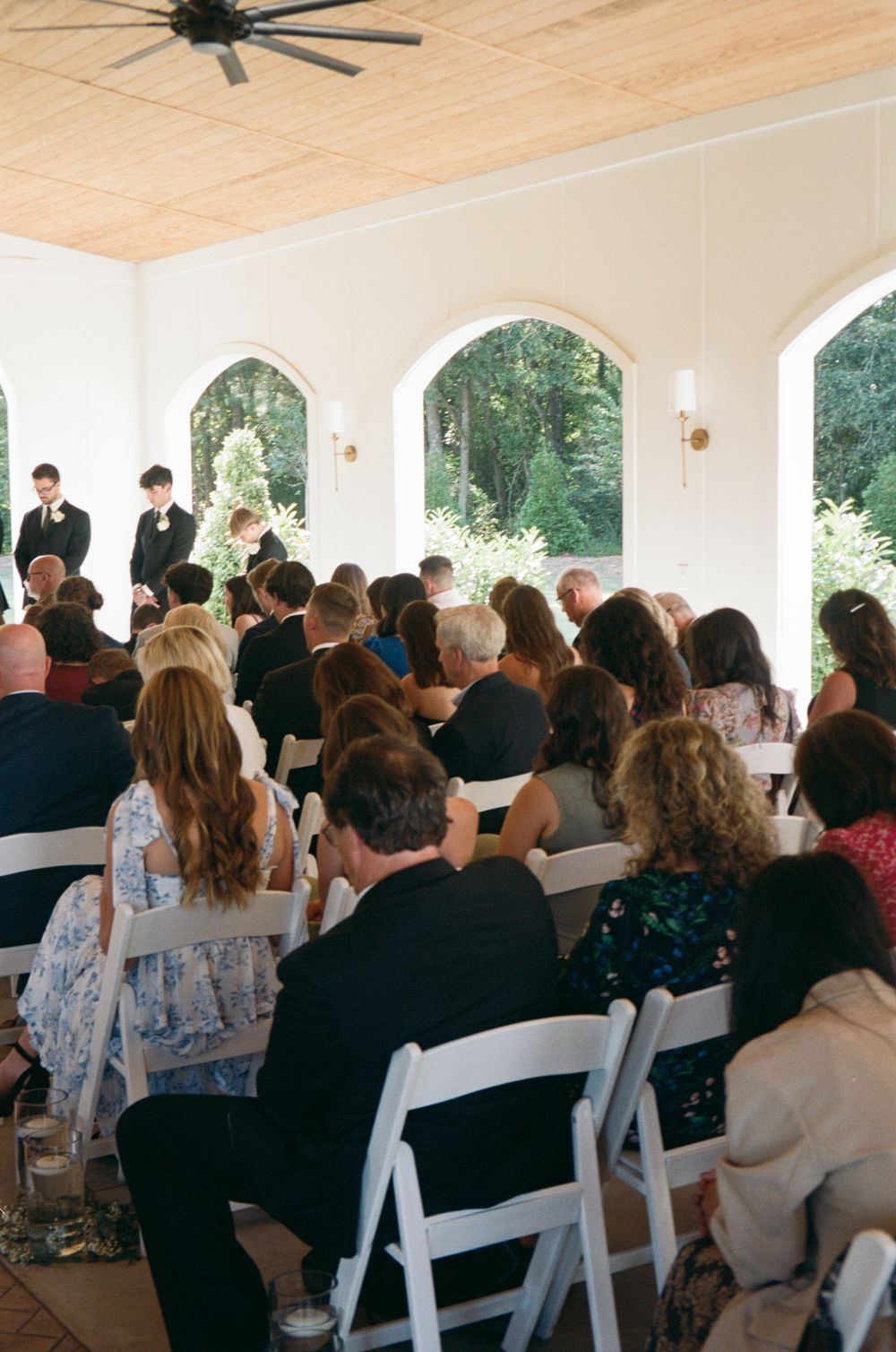 Wedding guests seated in white chairs during an outdoor covered ceremony with wooden ceiling beams and arches.