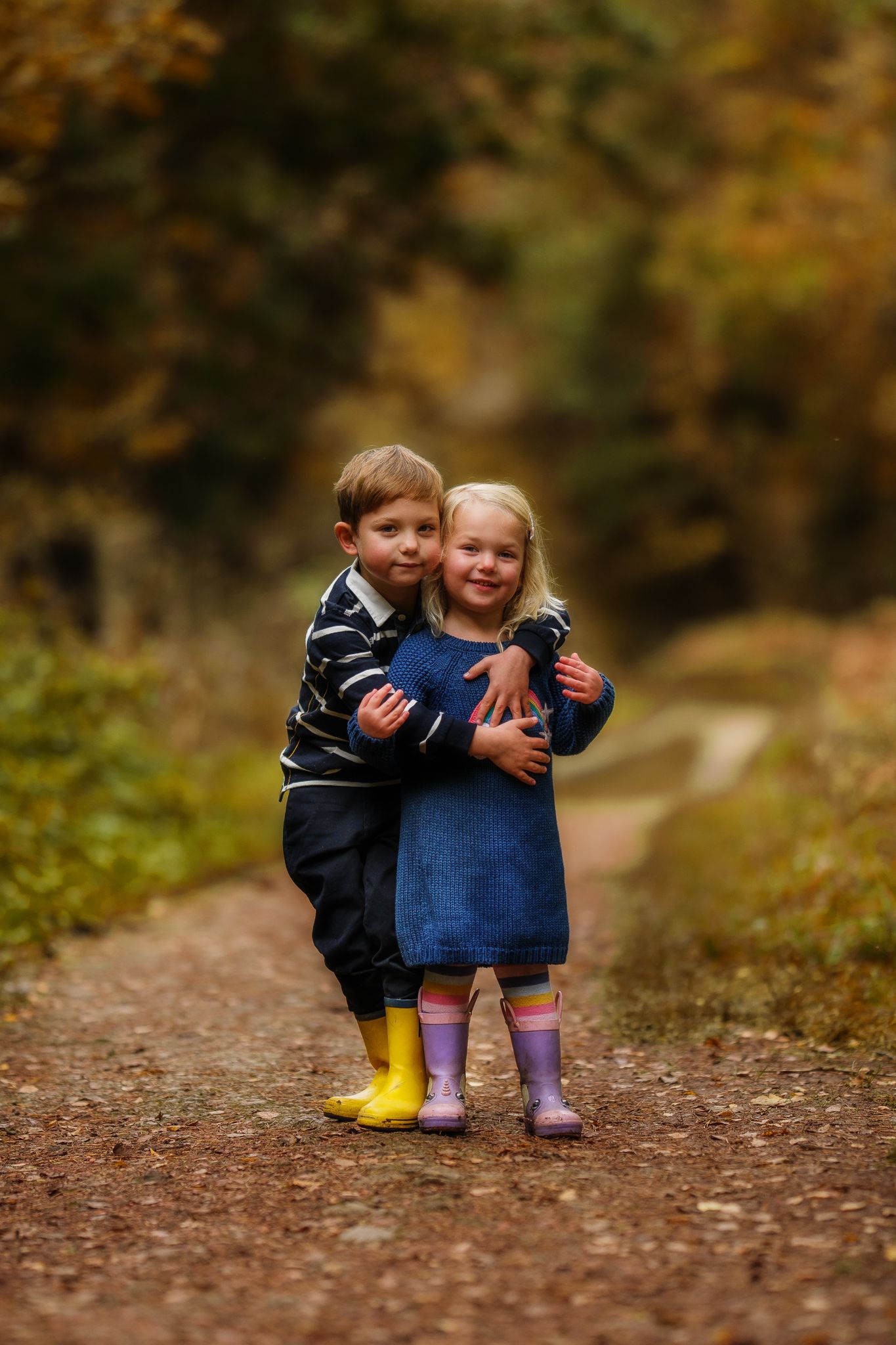 Two young children in winter coats share a sweet embrace on a forest path during autumn.