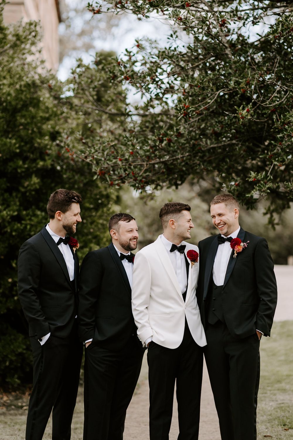 Groomsmen in black and white formal attire share candid laughs under tree branches at an outdoor wedding.