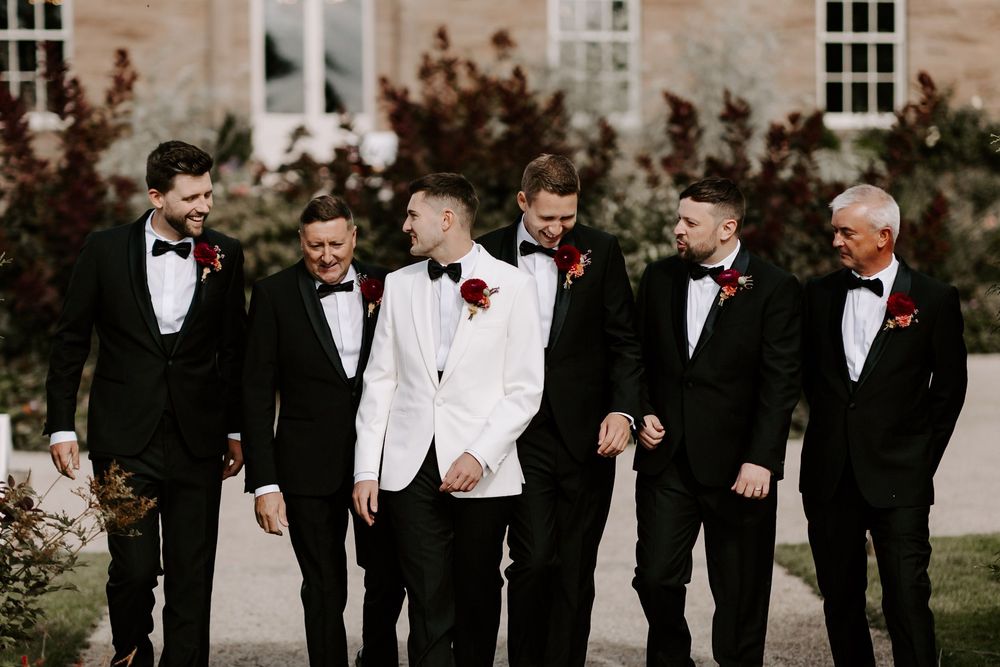 Wedding party in black tuxedos and red boutonnieres walk together and laugh outdoors.