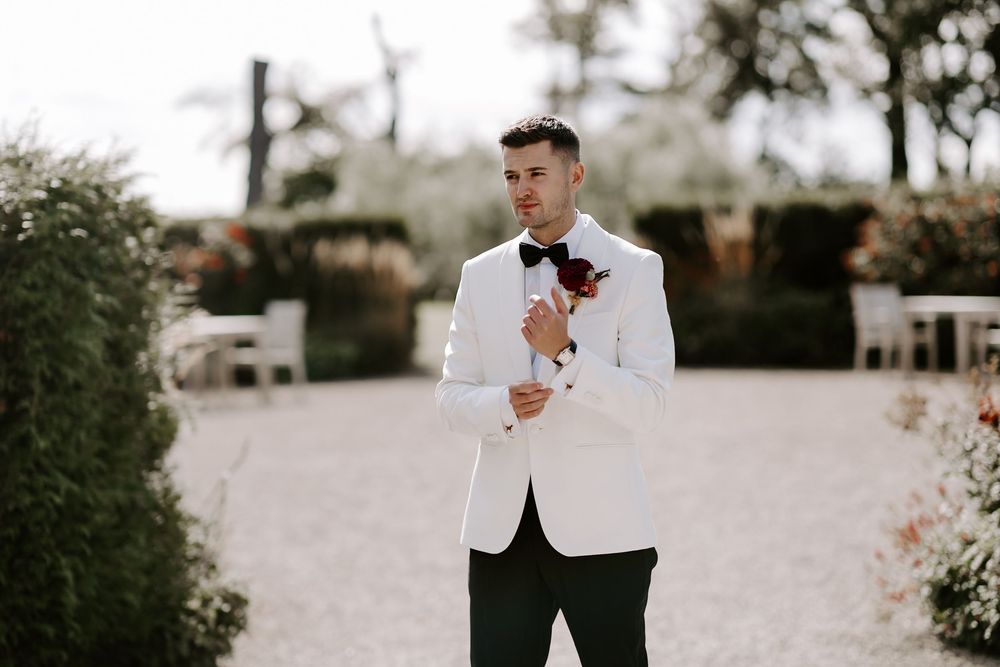 A person in a white tuxedo jacket holds a wine glass while standing outdoors at an elegant event.