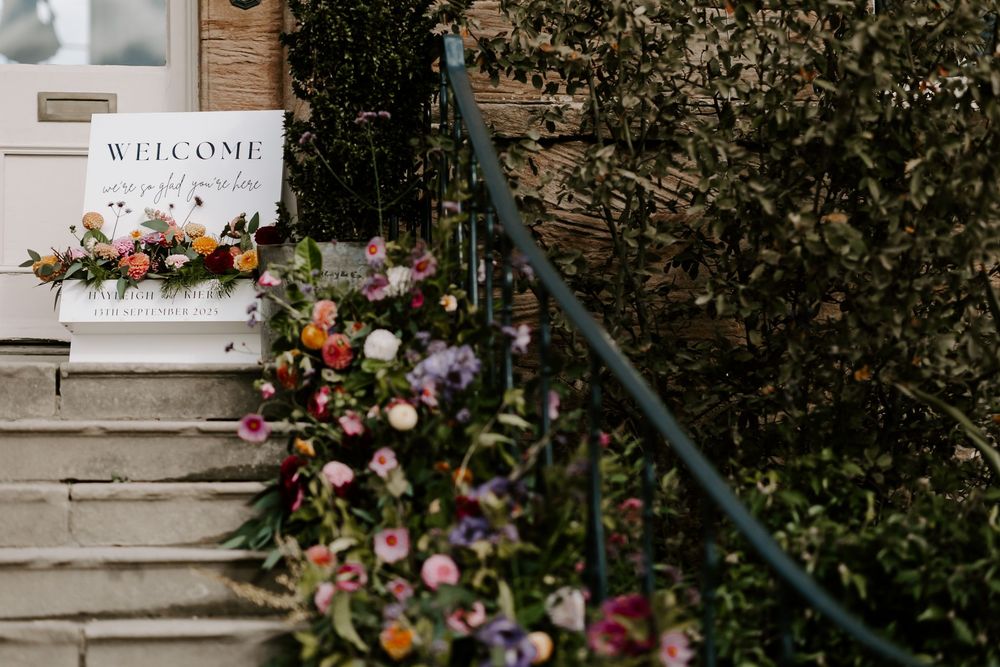 Elegant wedding welcome sign with floral arrangements displayed on a rustic stone wall.