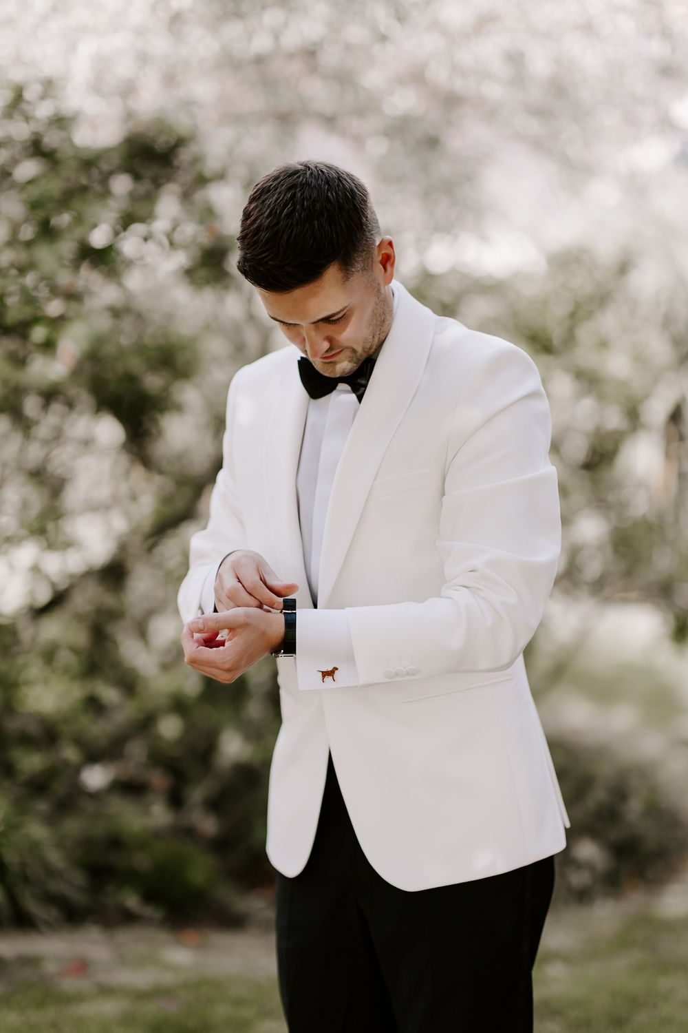 A groom in a white dinner jacket and black bowtie adjusts his cuff links during an outdoor wedding photoshoot.