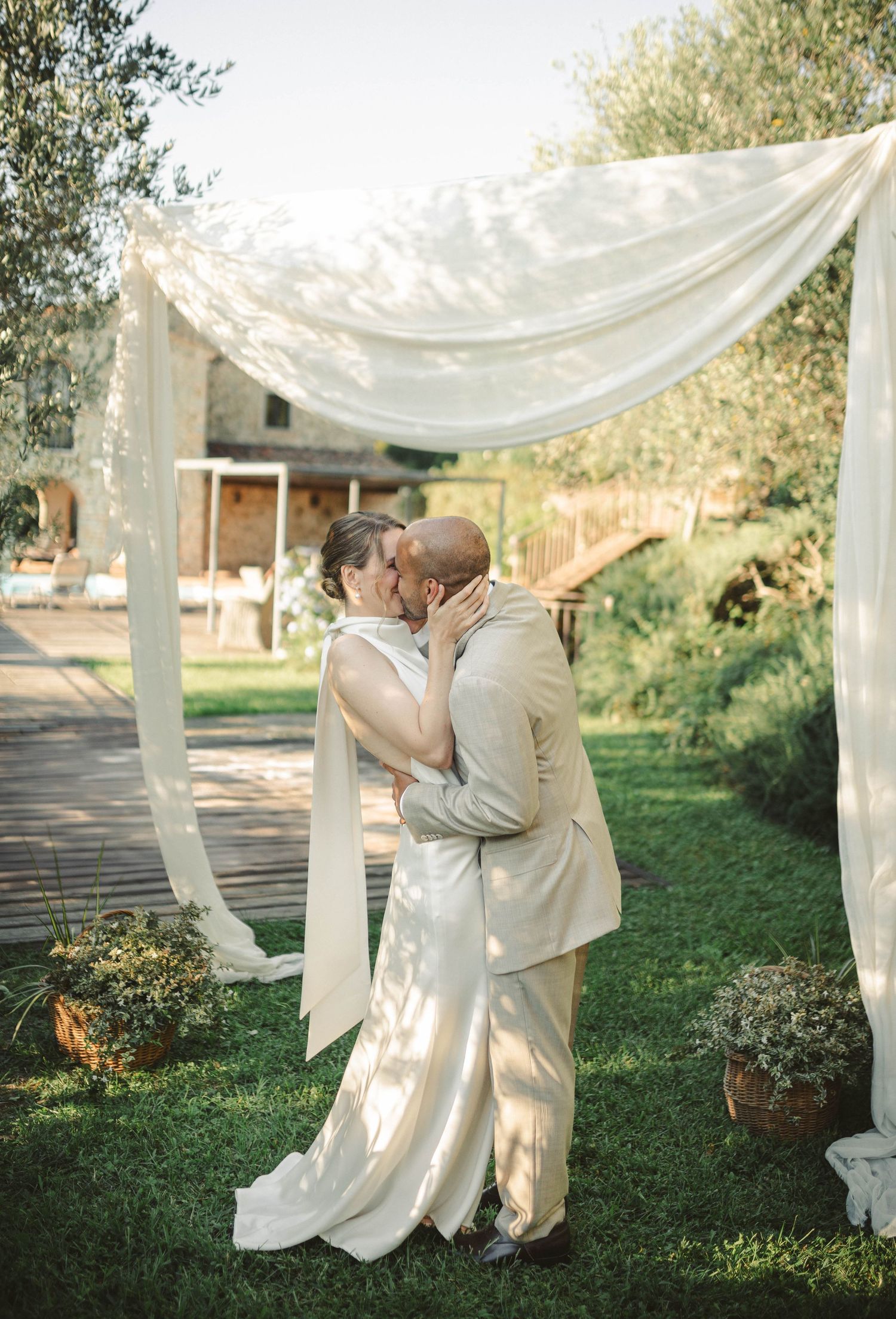 Newlyweds embrace under a flowing white fabric canopy in a garden setting.
