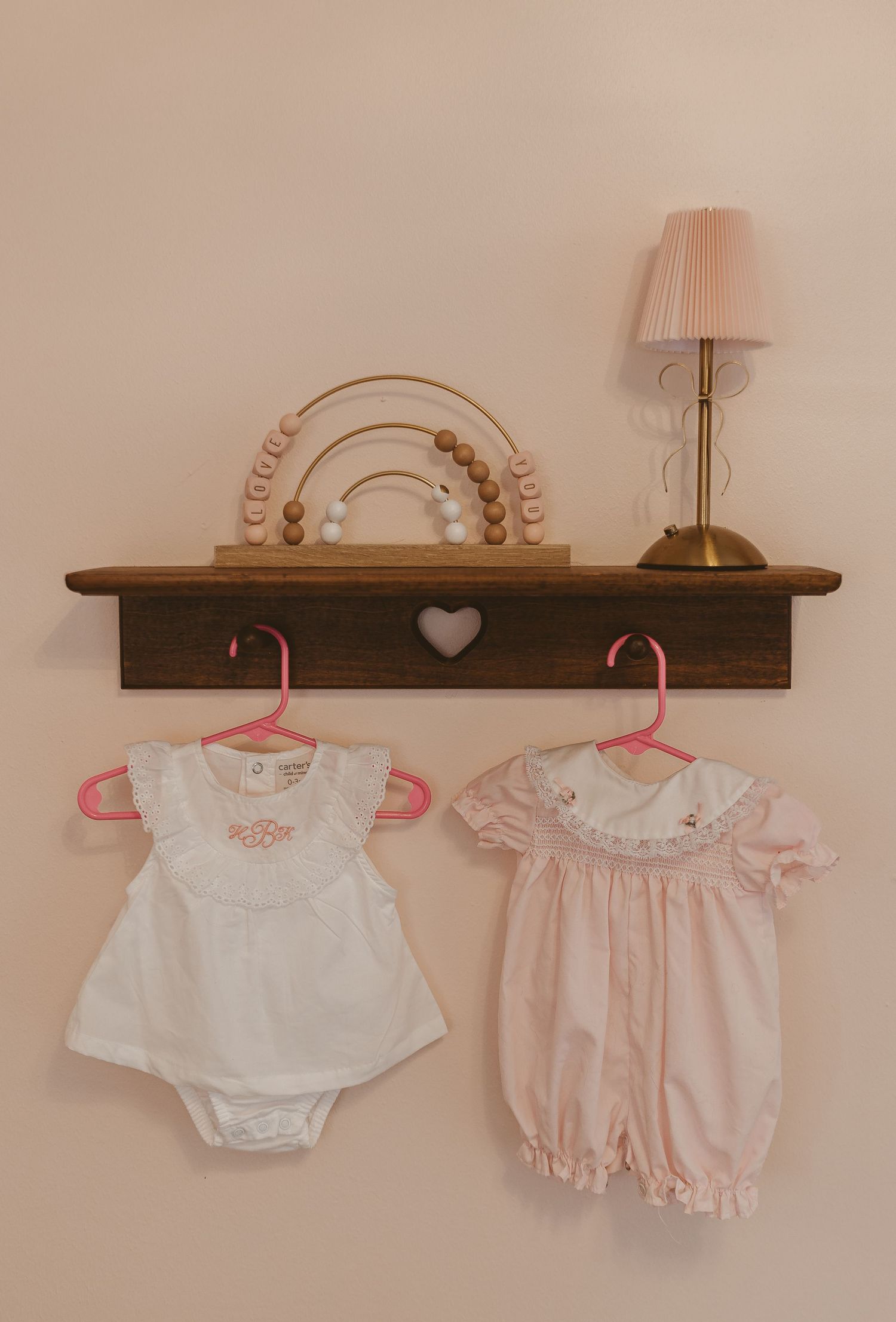 Baby clothes and onesies displayed on wooden wall shelf with pink hangers.