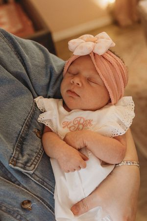 Sleeping newborn in white dress and pink bow headband resting on denim.