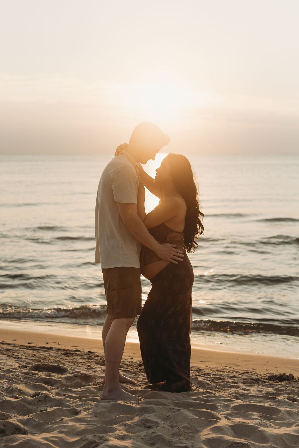 A couple embraces on a sandy beach at sunset with gentle waves rolling in the background.