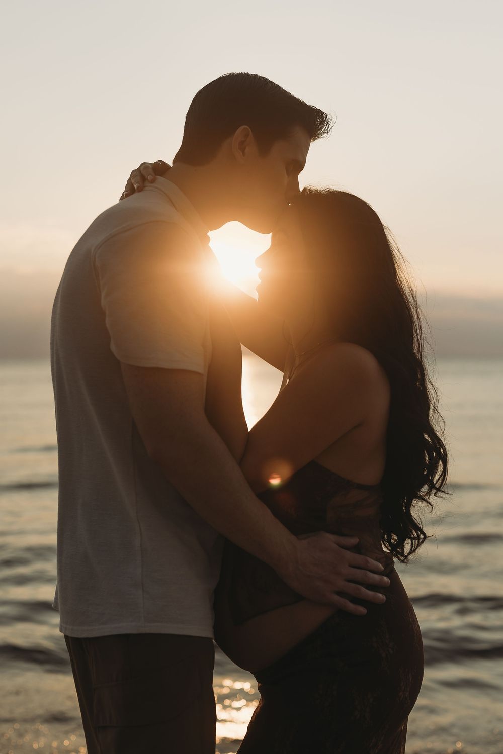 A silhouetted couple embraces on the beach at sunset with the sun glowing between them against an ocean backdrop.