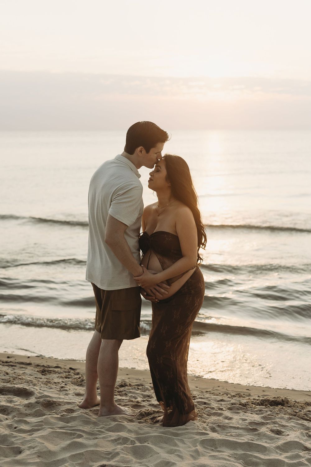 A couple shares an intimate moment on a sandy beach at sunset with ocean waves in the background.